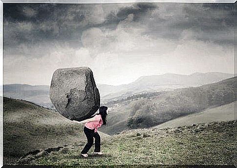 A woman is walking in a mountain landscape with a large rock on her back.